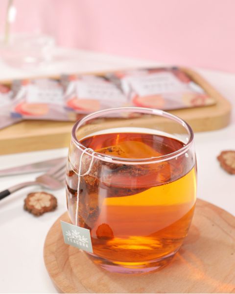 Glass cup with amber-colored herbal tea and tea bag, placed on a wooden saucer.