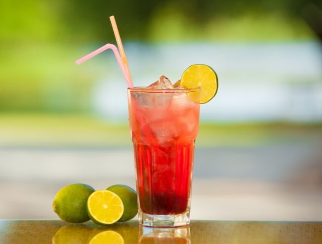 Glass of red non-alcoholic drink with ice cubes, lime slice and colorful straws, placed outdoors.