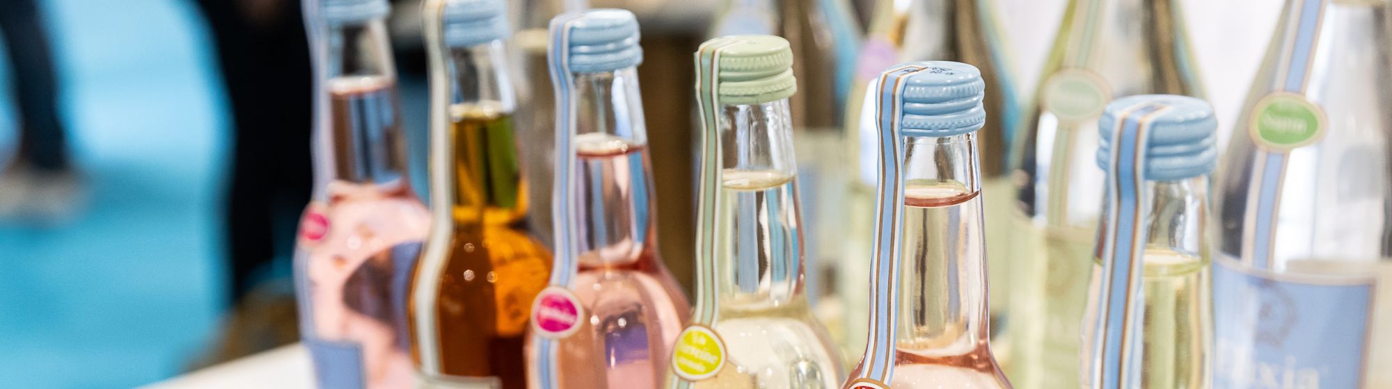 Row of glass bottles with colorful drinks and pastel caps, displayed on a booth.