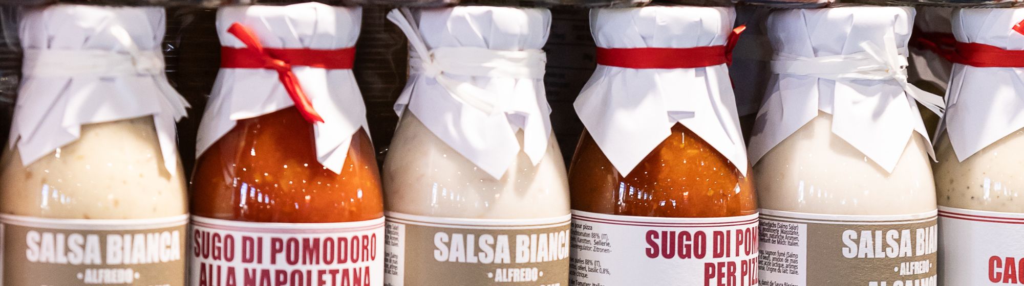 Jars of Italian white and red sauces lined up on a shelf, with paper-wrapped lids tied with string
