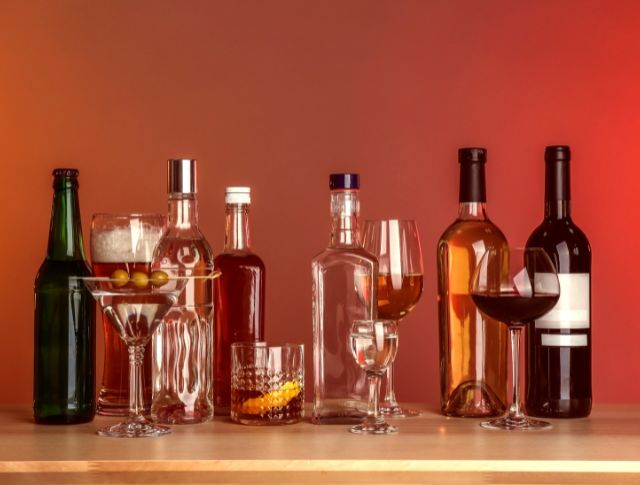 Assortment of alcoholic bottles and cocktail glasses on a wooden counter with a red background.