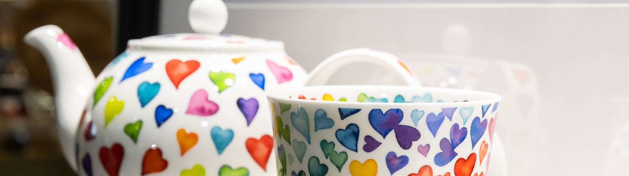 Théière et tasse blanches décorées de cœurs multicolores, posées sur une surface claire.