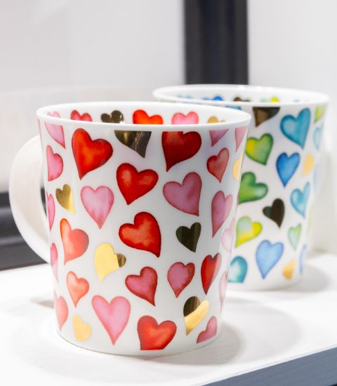 Deux mugs en porcelaine blanche décorés de cœurs multicolores et dorés, posés côte à côte sur une surface claire.