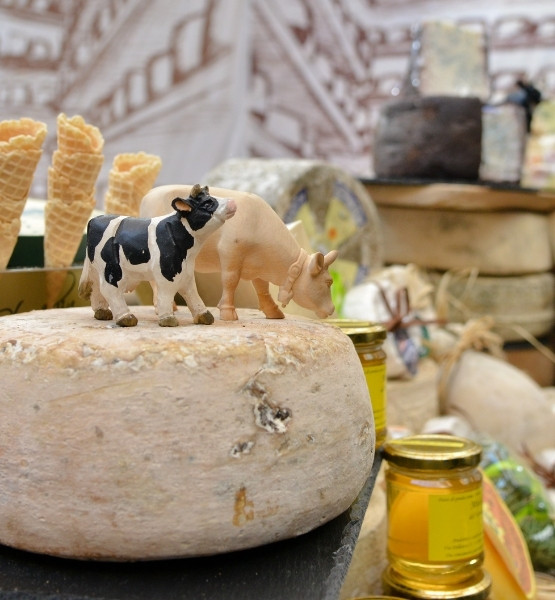 Cheese wheel topped with small cow figurines, honey jars and waffle cones in the background at a deli counter.