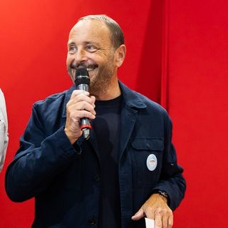 Homme souriant tenant un micro devant un fond rouge vif lors d’un événement ou d’une prise de parole.