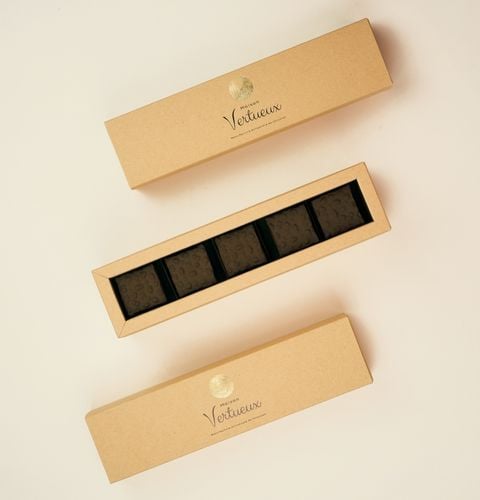Open beige gift box containing five dark chocolates, framed by two long closed boxes.