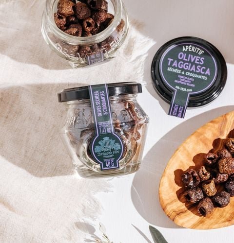 Jars of dried crispy Taggiasca olives, displayed with olives in a wooden bowl.