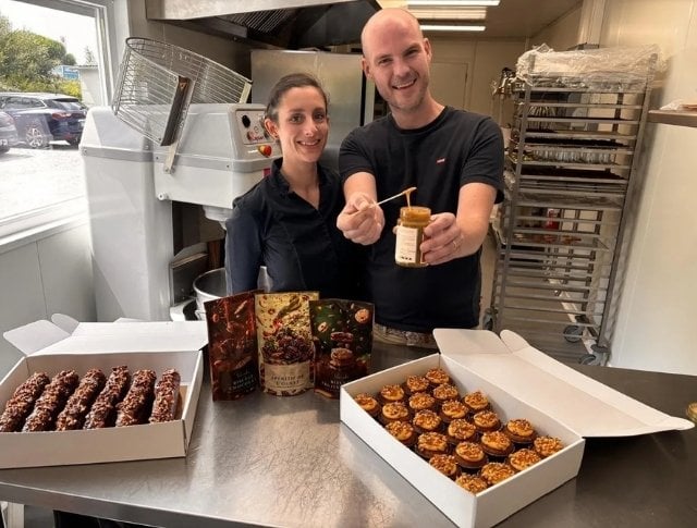 Two smiling pastry chefs presenting desserts and a jar of caramel in a professional kitchen